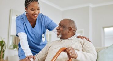 Smiling caregiver in blue scrubs supporting an elderly man with a cane at home