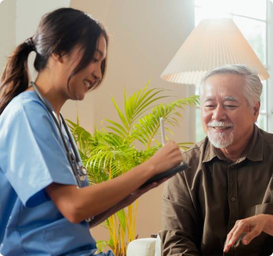 Smiling caregiver in blue scrubs taking notes on a tablet while chatting with a senior man at home
