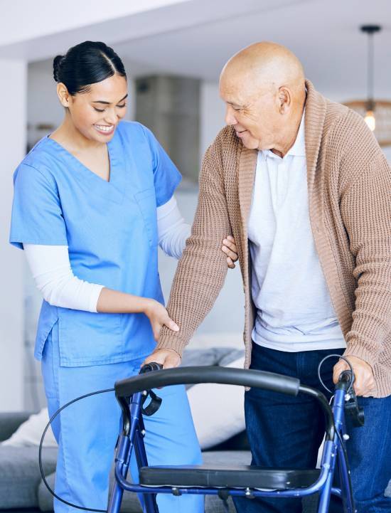 A nurse in blue scrubs gently assists an elderly man using a walker. The scene conveys warmth and care in a home setting, highlighting supportive healthcare.