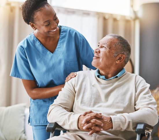 A smiling caregiver in blue scrubs gently rests her hand on the shoulder of an elderly man in a wheelchair. They share a warm, caring moment.