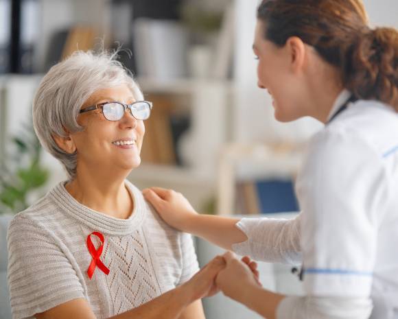 An elderly woman wearing glasses and a red ribbon smiles at a young nurse in a white uniform. They hold hands in a warmly lit room, conveying care and support.