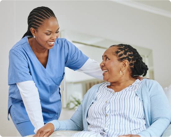 Caregiver in blue scrubs smiling and resting a hand on the shoulder of a laughing older woman sitting up in bed.
