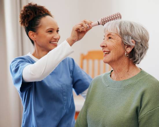 Flexible 24-Hour Care With No Long-Term Commitments in Edmonton -image A caregiver in blue scrubs cheerfully brushes the hair of an elderly woman with short gray hair, who is smiling, creating a warm, caring atmosphere.