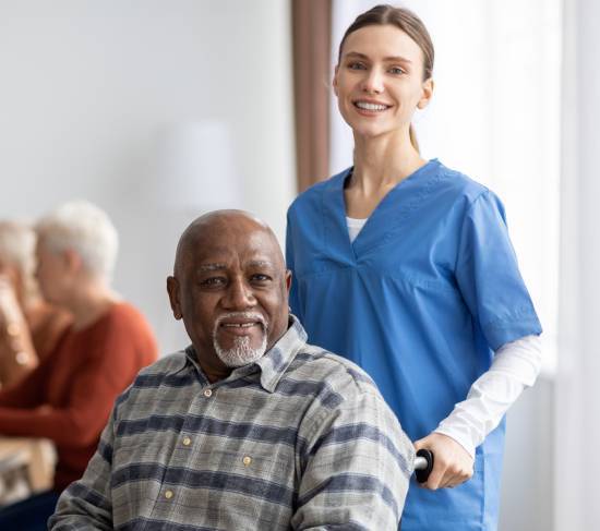 Getting Started with Parkinson_s Home Care Services in Edmonton-image A smiling nurse in blue scrubs stands beside an elderly man in a wheelchair, conveying warmth and care. Other seniors are visible in the background.