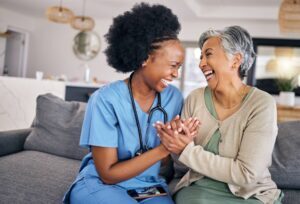 Caregiver in blue scrubs and senior woman with grey hair laughing together while holding hands on a couch in a bright living room