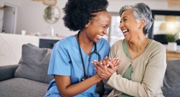 Home-Care-vs-Nursing-Home Caregiver in blue scrubs and senior woman with grey hair laughing together while holding hands on a couch in a bright living room