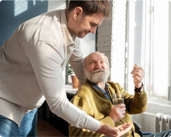 How Live-In Care Works-image A smiling elderly man sits by a window holding a glass of water. A younger man leans in, offering him a pill. The scene conveys care and warmth.