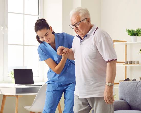 A nurse in blue scrubs kindly assists an elderly man in a white polo with walking. They are in a bright room with a window and laptop, conveying care and support.