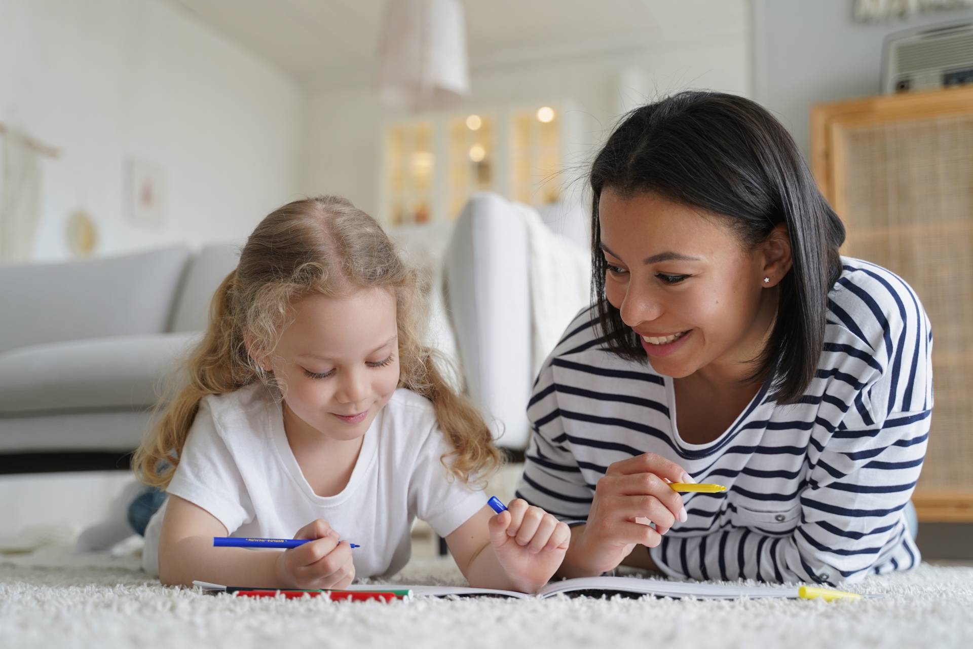 Woman and young girl lying on carpet together, drawing with colorful markers in a bright living room