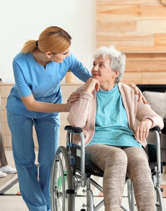 A nurse in blue scrubs compassionately smiles at an elderly woman in a wheelchair, gently touching her shoulder in a cozy, warmly lit room.