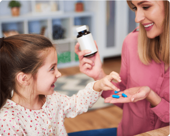 Smiling woman holding a supplement bottle and offering blue vitamins to a young girl in a home setting