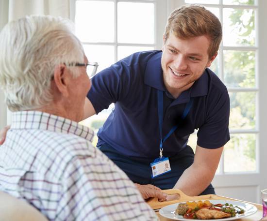 A caregiver in a blue uniform smiles warmly at an elderly man with white hair and glasses, who is seated at a table with a meal in a well-lit room.