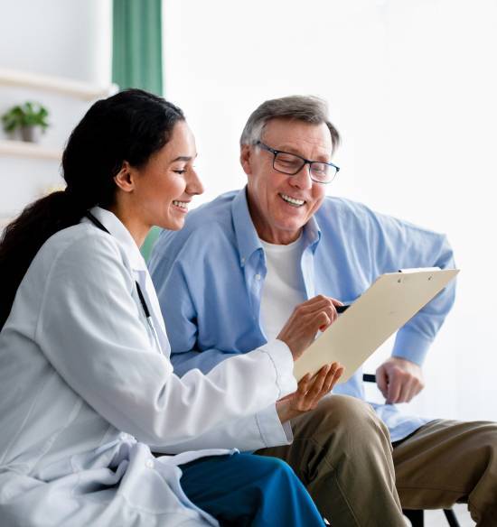 A smiling doctor explains medical notes on a clipboard to an elderly patient. Both appear content and engaged in a bright, friendly office setting.