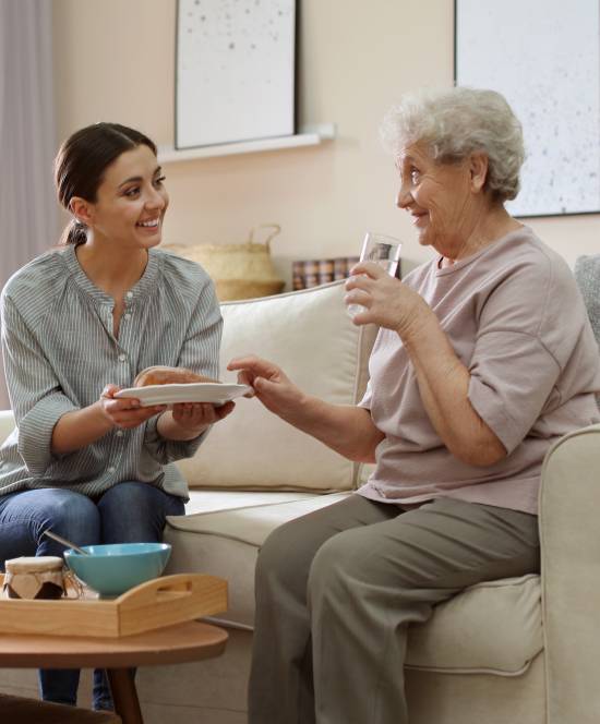 A young woman smiling, handing a plate to an elderly woman holding a glass of water, sitting on a sofa, conveying warmth and care in a cozy living room.