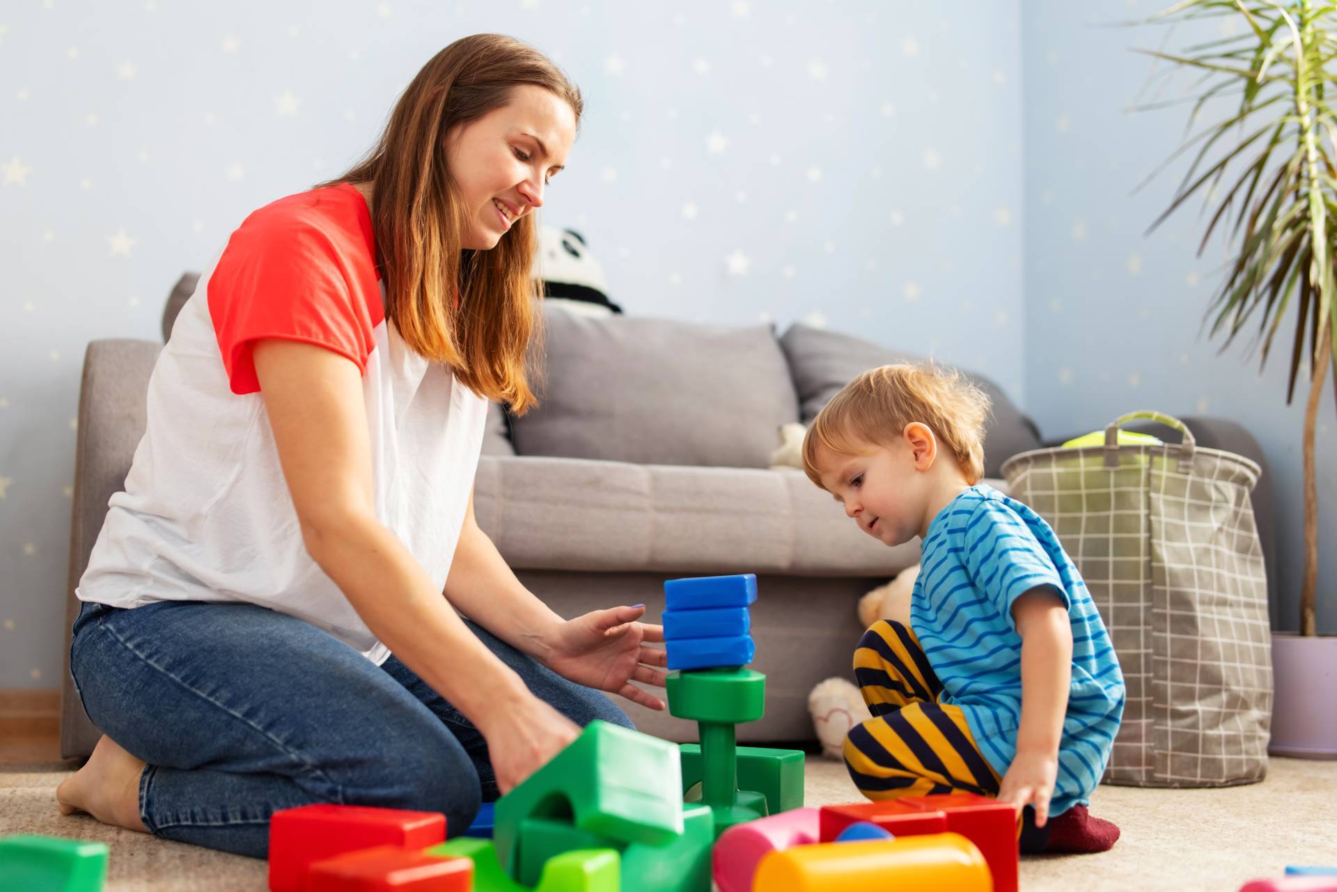 Woman and toddler sitting on the floor together building a tower with colorful wooden blocks in a cozy playroom