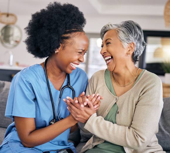 A nurse in blue scrubs shares a heartfelt laugh with an elderly woman on a couch. They hold hands warmly, conveying joy and connection in a light-filled room.
