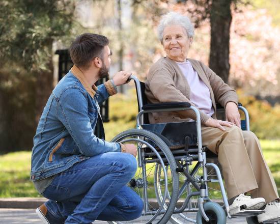A young man kneels beside an elderly woman in a wheelchair in a park. They smile warmly at each other, surrounded by greenery and soft sunlight.