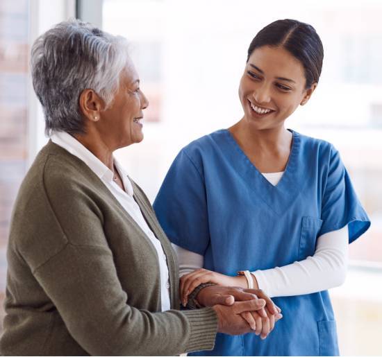 The Trusted Choice for 24-Hour Senior Home Care in Edmonton-image A caregiver in blue scrubs warmly holds an elderly woman's hands, both smiling. The scene conveys care and trust in a bright, peaceful setting.