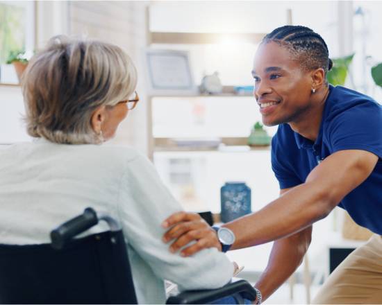 Three 8-Hour Shifts-image A smiling caregiver in a blue shirt leans forward while gently holding the arm of an elderly person in a wheelchair, creating a warm, supportive atmosphere.
