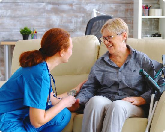 Home care nurse with a stethoscope kneeling beside a smiling senior woman seated on a couch, with forearm crutches nearby.