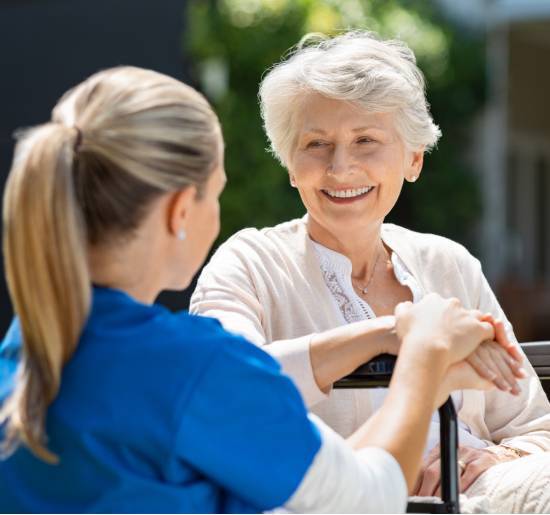 Two 12-Hour Shifts-image A smiling elderly woman and a caregiver share a warm conversation outdoors. The elderly woman sits in a wheelchair, conveying comfort and companionship.