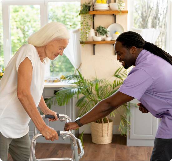 Two 12-Hour Shifts-image Caregiver in lavender scrubs guiding an elderly woman with a rolling walker across a bright, plant-filled kitchen.