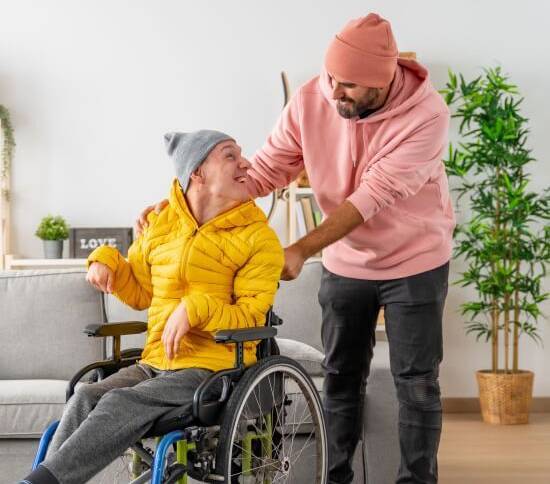 A man in a yellow jacket and gray beanie sits in a wheelchair, smiling at another man in a pink hoodie and beanie. The scene is warm and friendly, set in a cozy living room.
