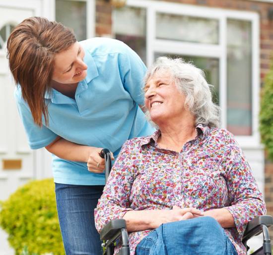 A smiling elderly woman in a wheelchair interacts warmly with a younger caregiver in a blue shirt. They're outside a house, conveying care and companionship.