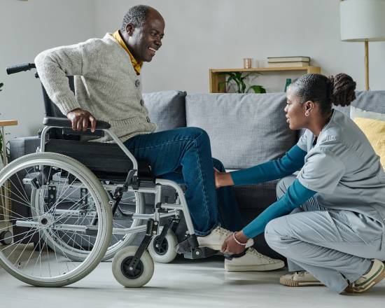 What Do Ohana Care_s 24-Hour Home Care Services in Edmonton Include_-image A caregiver in scrubs assists a smiling man in a wheelchair, adjusting his footrest. The room is bright and cozy, featuring a couch, plants, and books.