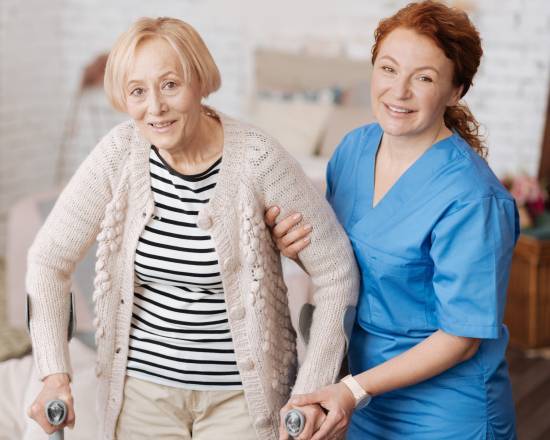 Elderly woman with a walker is assisted by a smiling caregiver in blue scrubs. The setting is warm and supportive, suggesting a caring environment.