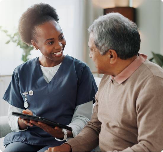 What Is Elder Live-In Care-image Caregiver in navy scrubs holding a tablet and smiling while talking with a senior man seated on a couch.
