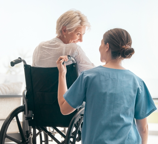 Caregiver pushing an elderly woman in a wheelchair by a bright window