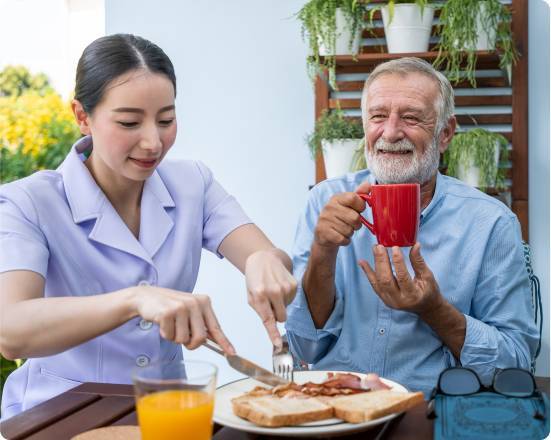 Caregiver cutting food for a smiling senior man holding a mug of coffee at an outdoor breakfast table.