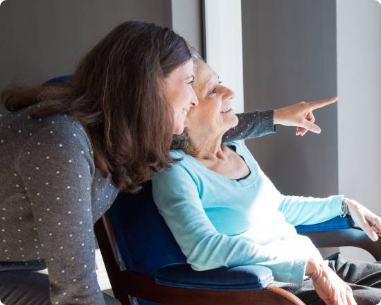 Caregiver and senior woman sitting by a window, smiling and pointing outside together