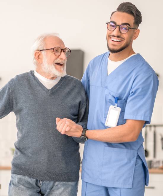 A smiling elderly man in glasses holds hands with a young male nurse in blue scrubs. They both appear happy, conveying a sense of care and support.