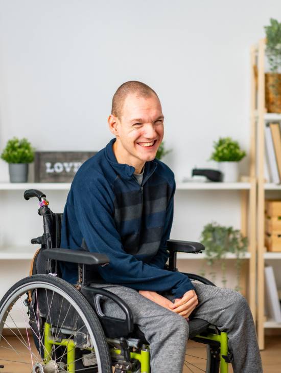 A person in a wheelchair smiles warmly in a cozy room with plants and shelves. They wear a striped sweater and gray pants, conveying a joyful and inclusive atmosphere.