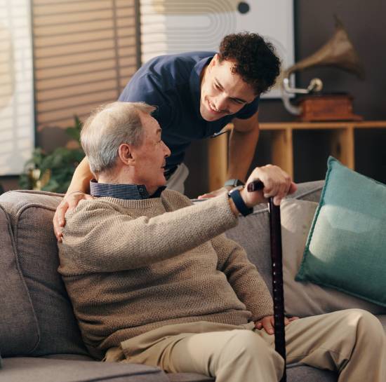 Your Trusted Provider For Parkinson_s Home Care Services in Edmonton-image A young man gently supports an elderly man with a cane on a sofa. They share a warm smile, conveying companionship and care in a cozy living room.