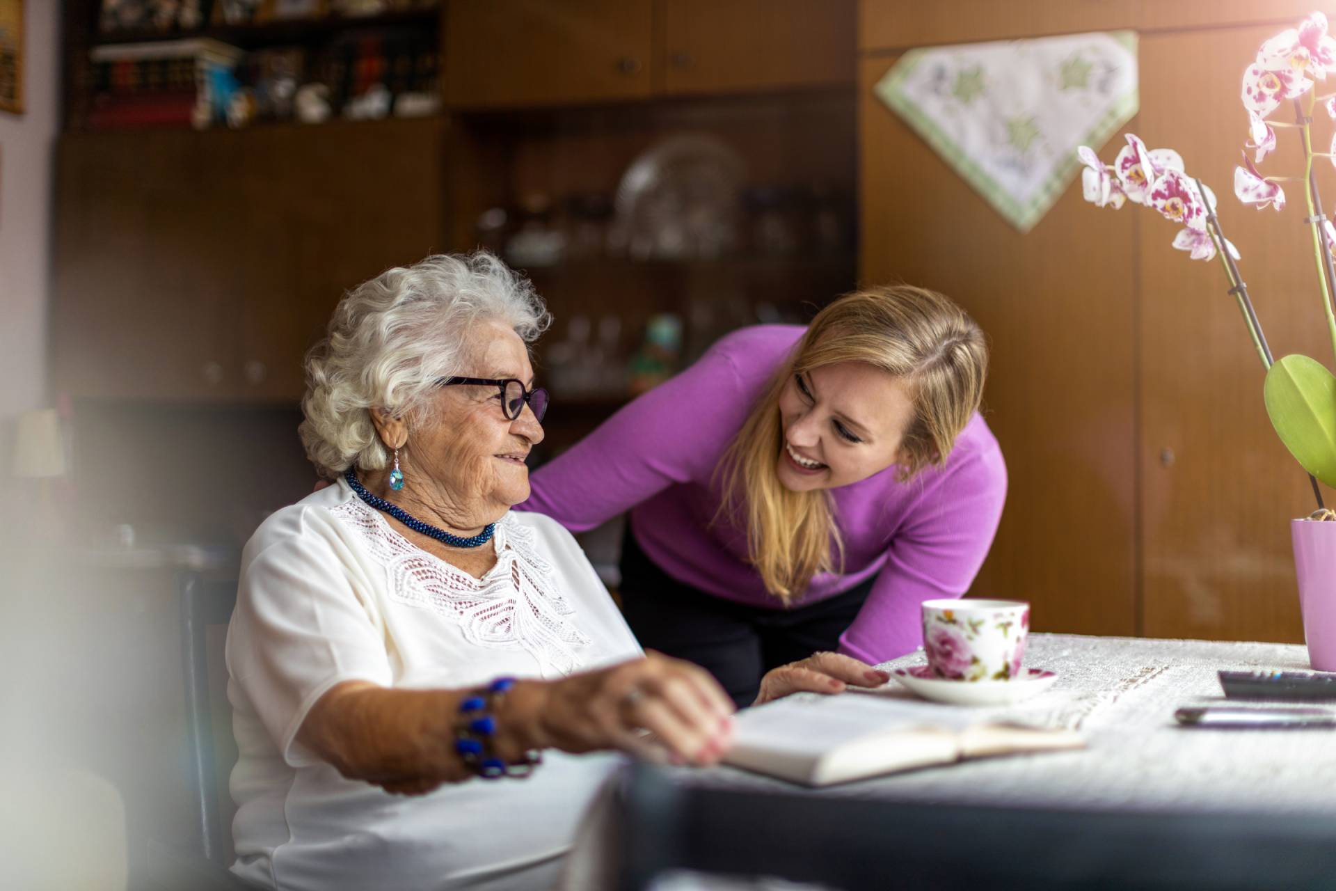 Elderly woman with white hair and glasses smiles at a younger woman in a purple sweater. They're at a table with a book and teacup, in a cozy room.