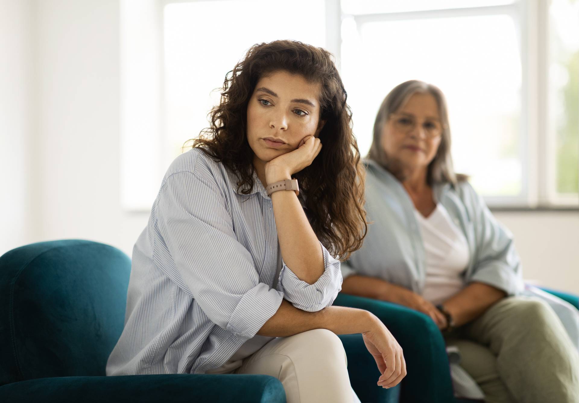 Young woman sitting with her chin resting on her hand, looking thoughtful while an older woman sits in the background.
