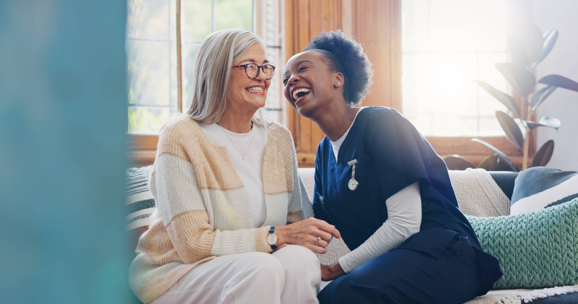 Smiling nurse in navy scrubs sitting on a couch and laughing with an elderly woman at home.