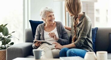 n older woman and a younger woman sitting together on a couch, smiling and talking while looking at a tablet. The setting appears to be a bright, cozy living room with natural light, blue pillows, and a coffee table with mugs and books.
