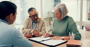 An older couple sitting at a table reviewing documents with a financial advisor. The man is pointing to a section on a paper while the woman holds a pen, listening attentively. They appear to be in a bright office or home setting with notebooks and pens on the table.