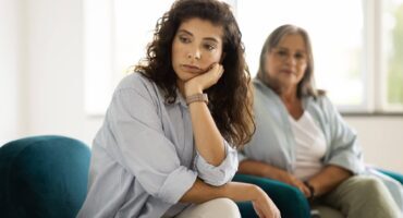 Young woman sitting with her chin resting on her hand, looking thoughtful while an older woman sits in the background.