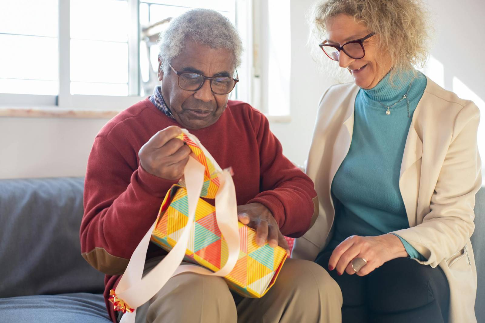 An elderly man in a red sweater, with glasses, opening a gift with colourful wrapping paper, while a smiling woman sits beside him.