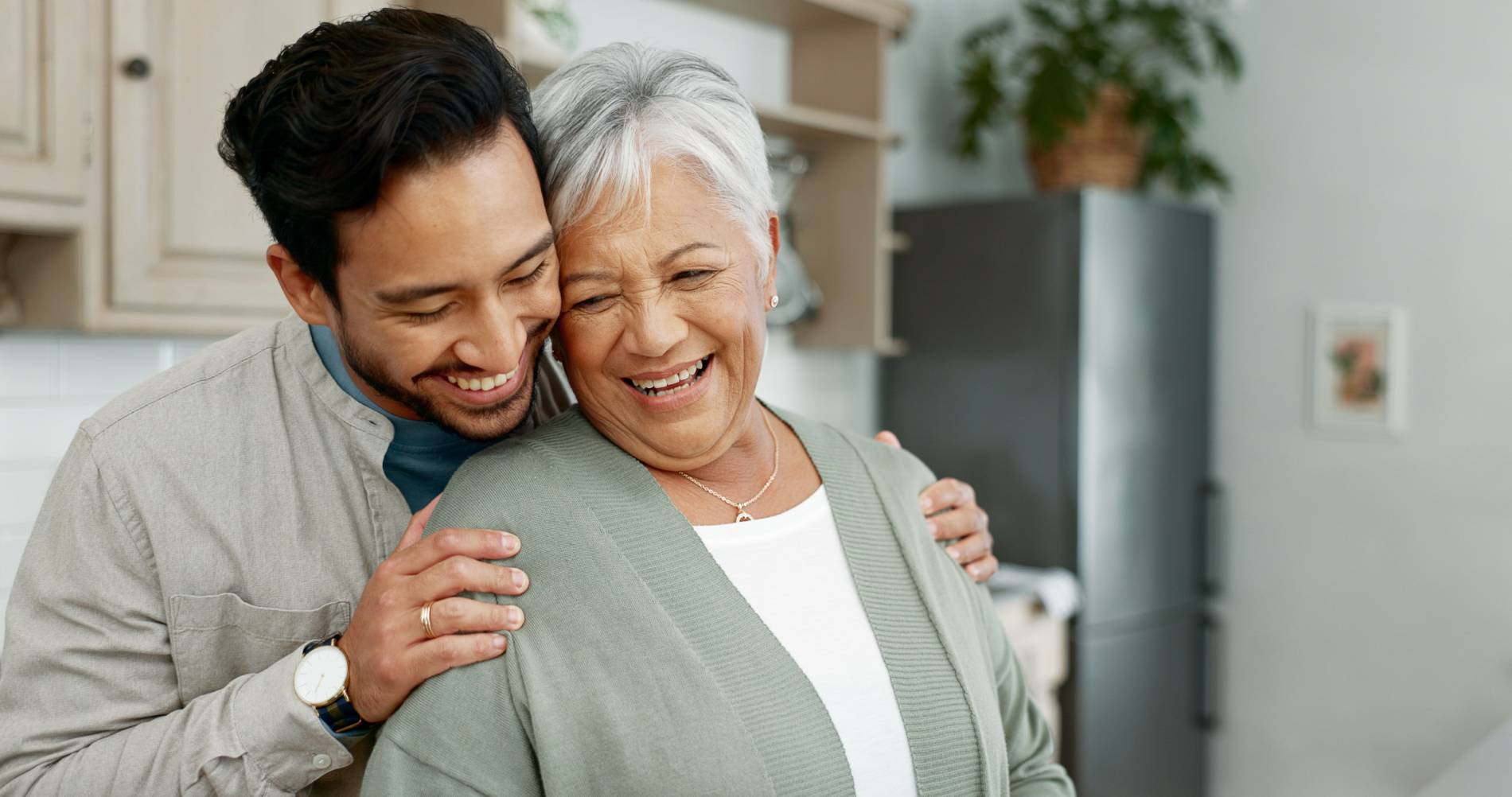 Smiling adult son embracing his elderly mother in a warm hug in a cozy kitchen setting.