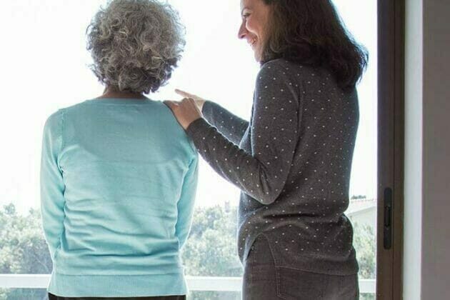 memory-care-edmonton Elderly woman and her caregiver standing and looking out of a window.
